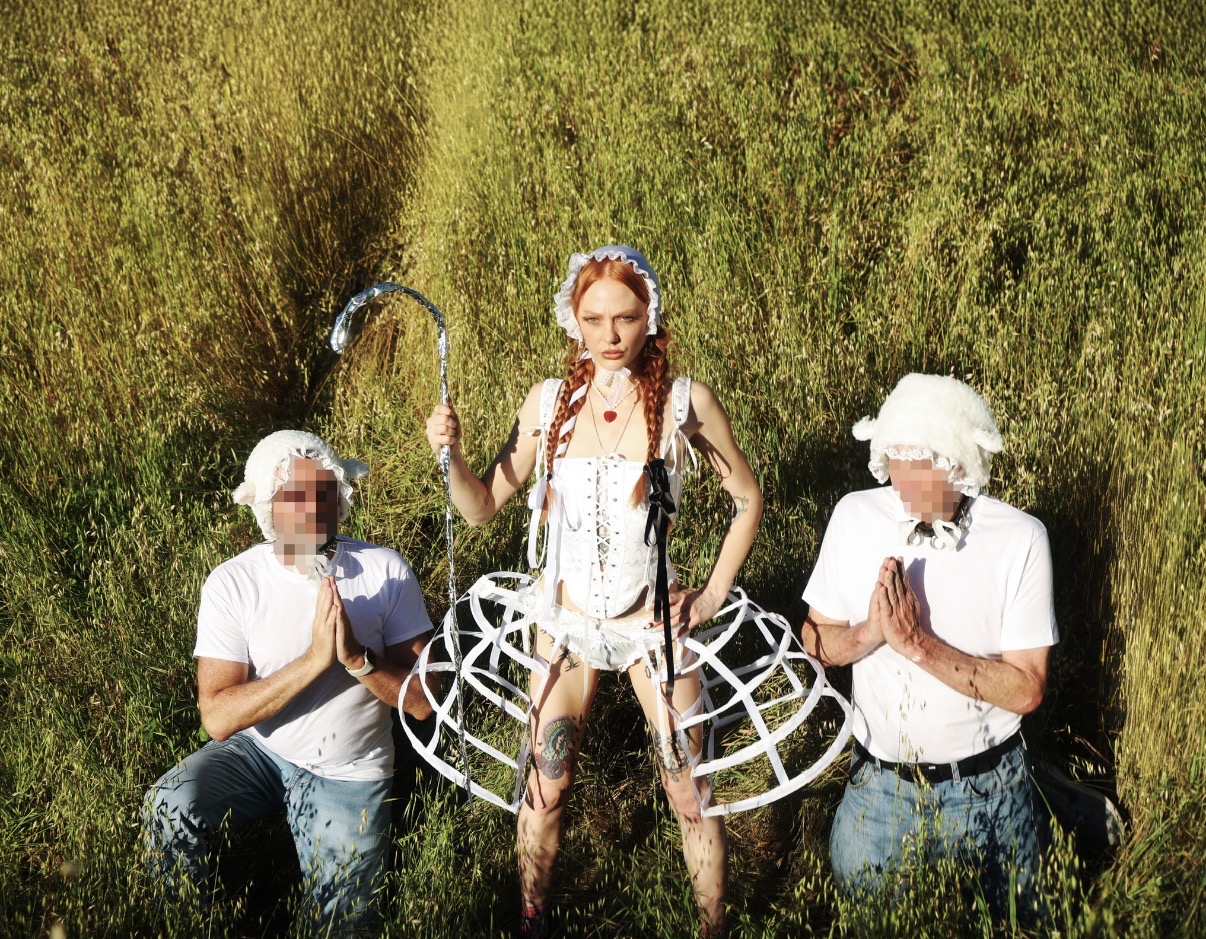 Mistress Luna in white shepherdess styling framed by two kneeling figures in a devotional gesture